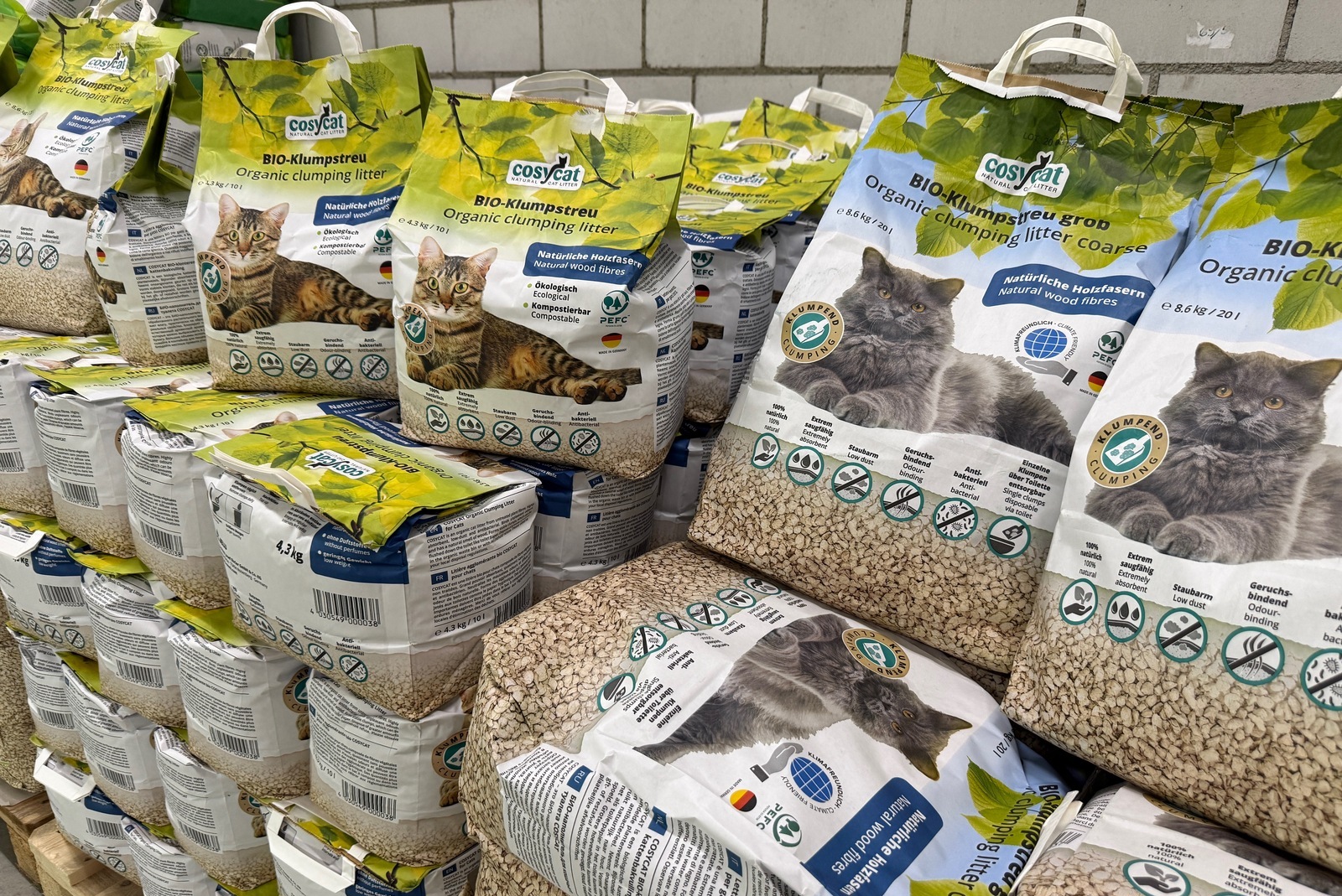 Stacks of green-and-white bags labeled 'BIO-Klum pstreu Organic clumping litter' with cat images on each bag, piled against a cinder block wall, ready for sale.